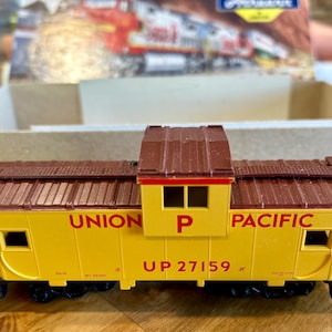 May include: Yellow Union Pacific caboose model with brown roof and details. The words "UNION PACIFIC" and "UP 27159" are printed on the side. The model has black wheels and railings.