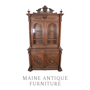 May include: A tall, dark brown wooden cabinet featuring two glass-paned upper doors, two lower solid doors with carved bird motifs, and barley-twist columns on the sides. The top is adorned with an ornate pediment and finials. Text reads: MAINE ANTIQUE FURNITURE.