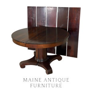 May include: A dark brown wooden round pedestal table with a split top and a central column base featuring four curved feet. Behind the table stand five vertical wooden panels of the same dark brown finish. The text MAINE ANTIQUE FURNITURE is below.