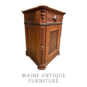 May include: A wooden bedside cabinet featuring a light-colored stone top, one drawer with a decorative carved handle, and a single door with a recessed panel. The piece has dark wood accents, turned corner details, and a sturdy base structure.