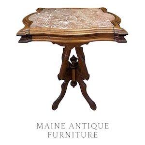 May include: A wooden pedestal table featuring a square top with scalloped edges and a brown marble inlay with white veining. The base consists of a central column supported by four curved legs, with the text MAINE ANTIQUE FURNITURE below.
