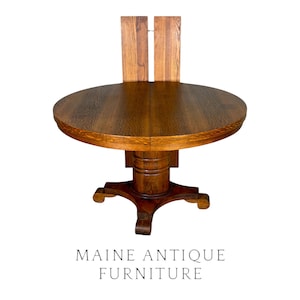 May include: A round wooden dining table with a dark brown finish and visible grain patterns. The table features a central pedestal base with four feet and two vertical wooden panels extending upward from the back of the tabletop. Text reads MAINE ANTIQUE FURNITURE.