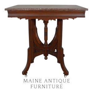 May include: A rectangular wooden table with a mottled brown marble top. The base features a central turned column flanked by curved supports and decorative carved trim. The table stands on four legs equipped with small white casters. MAINE ANTIQUE FURNITURE.