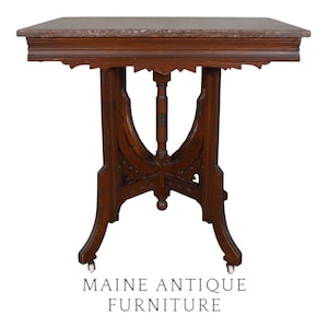 May include: Antique wooden side table with a rectangular marble top. The table has ornate carved details on the legs and apron, and is supported by four legs with small wheels. The text "MAINE ANTIQUE FURNITURE" is at the bottom.