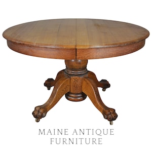 May include: A round wooden dining table with a smooth top and a central pedestal base. The base features four curved legs ending in carved lion paw feet with small metal casters. The wood has a medium brown finish with visible grain patterns.