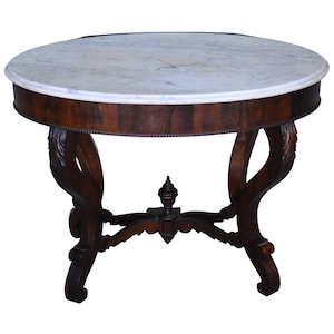 May include: A round table featuring a white marble top with grey veining and a dark brown wooden base. The table has a curved apron with a beaded edge, four ornate curved legs, and a central finial connecting the legs with a cross stretcher.