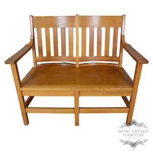 May include: A medium-brown oak wood bench featuring a slatted backrest, flat armrests, and a solid seat. The bench has a central vertical support beam and four legs connected by horizontal stretchers. A logo reads Maine Antique Furniture Est 1970.