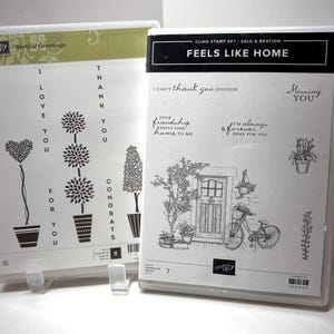 May include: Two clear plastic cases containing cling stamp sets. The left case is labeled Vertical Greetings and features vertical text and potted plant designs. The right case is labeled FEELS LIKE HOME and features a bicycle and house illustration.