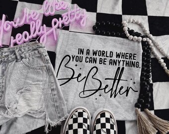 Peut inclure: Un t-shirt gris avec le texte "Dans un monde où vous pouvez être n'importe quoi, Soyez meilleur." Le t-shirt est posé sur une surface à carreaux noirs et blancs avec un collier de perles noires et blanches et une paire de Vans à carreaux noirs et blancs.