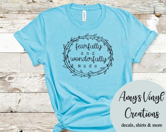 Peut inclure: Un t-shirt bleu clair avec un motif noir qui dit "fearfully and wonderfully made" à l'intérieur d'une couronne. Le motif est centré sur le t-shirt.