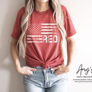 May include: Red t-shirt featuring a distressed American flag design with the word "RED" in white. The shirt has a crew neck and short sleeves. The text "REUNITED EVERYONE DEPLOYED" is also visible. The shirt is tied at the waist.