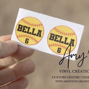 May include: Two yellow and orange softball stickers with black stitching and the name "Bella" and the number "6" printed on them. The stickers are on a white background with the text "Amy's Vinyl Creations" and "Custom Graphic Tees & More" printed below.