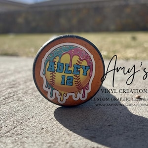May include: A baseball bat end cap with a custom design. The design features the name "ADLEY" and the number "12" in a teal font, with a baseball and a donut-like border in pink and blue. The cap is orange with black trim.