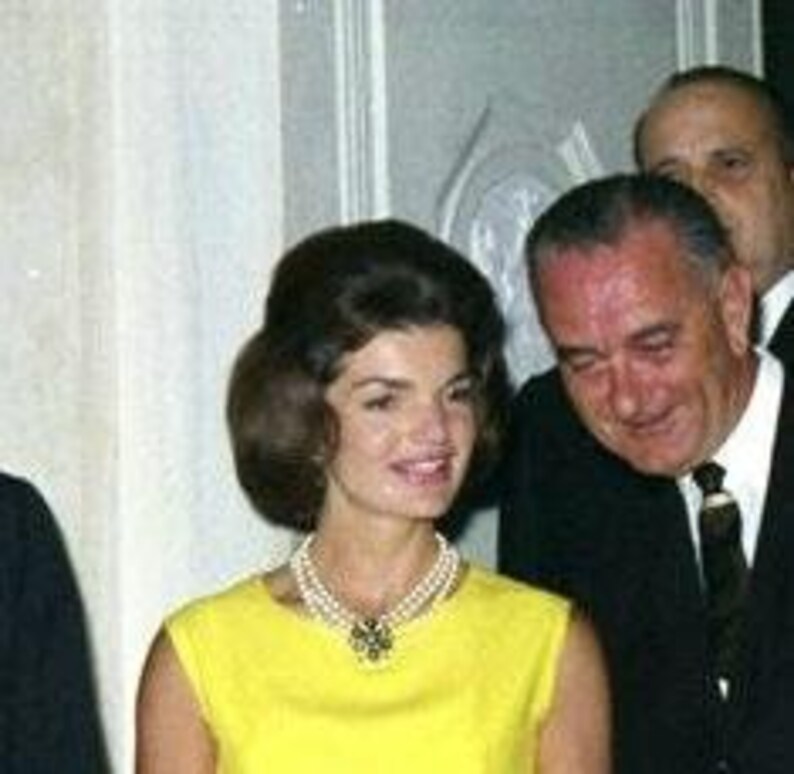 May include: A woman in a yellow dress with a pearl necklace and a brooch stands next to a man in a suit and tie. The woman is smiling and the man is looking down at her.