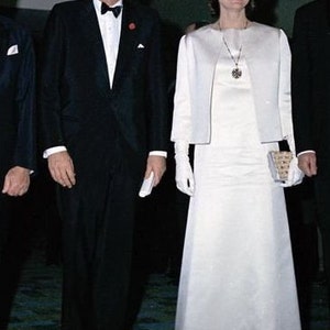 May include: A man and a woman in formal attire. The man is wearing a black tuxedo with a white shirt and a black bow tie. The woman is wearing a white dress with a white jacket and white gloves. They are standing on a patterned carpet.