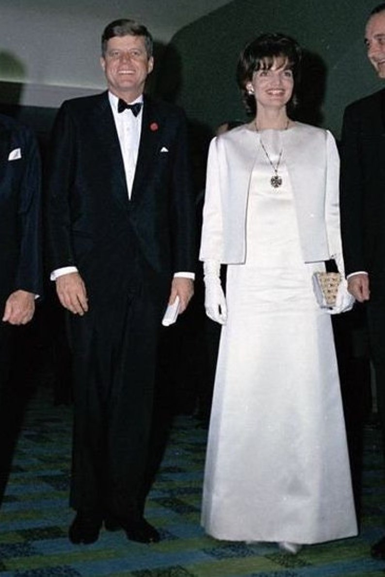May include: A man and a woman in formal attire. The man is wearing a black tuxedo with a white shirt and a black bow tie. The woman is wearing a white dress with a white jacket and white gloves. They are standing on a patterned carpet.