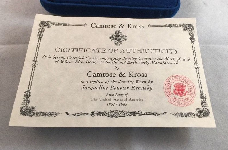 May include: A certificate of authenticity for jewelry, stating that the piece is a replica of the jewelry worn by Jacqueline Bouvier Kennedy, First Lady of the United States of America from 1961 to 1963. The certificate is signed by Camrose & Kross and features a seal of the President of the United States.