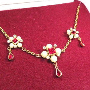 May include: A gold-tone necklace featuring three floral-inspired stations, each centered with a square-cut red stone surrounded by four round white beads and small clear crystals. Two teardrop-shaped red stones hang from the chain links.
