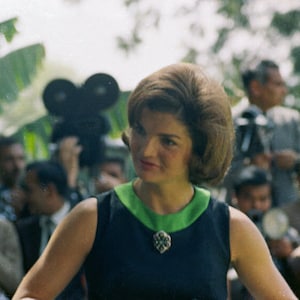 May include: A woman with dark hair styled in a bouffant wears a dark blue sleeveless dress with a green collar and a silver brooch. She is surrounded by photographers.