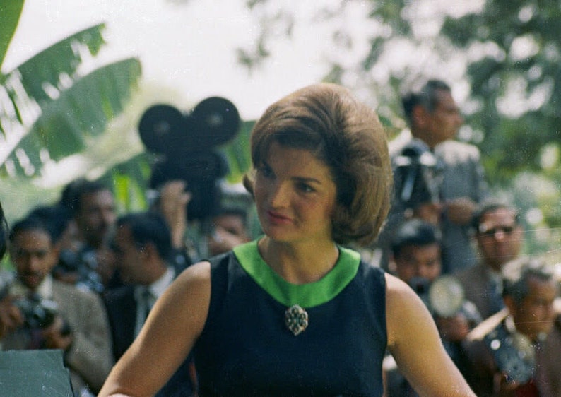 May include: A woman with dark hair styled in a bouffant wears a dark blue sleeveless dress with a green collar and a silver brooch. She is surrounded by photographers.