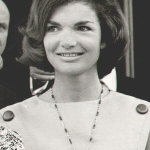 May include: A woman with short, dark hair is wearing a white collared shirt with a silver chain necklace and a large, ornate pendant. The pendant is a silver flower with black gemstones.