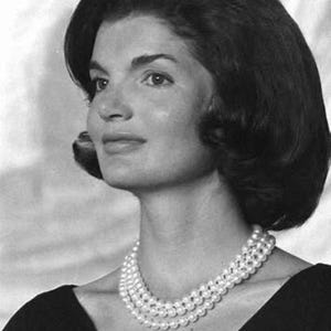 May include: Black and white portrait of a woman with dark hair and a three-strand pearl necklace. The woman is looking slightly to the left, with a soft focus background. She is wearing a black top.