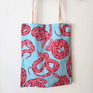 May include: A light blue tote bag with a repeating pattern of pink snakes. The snakes have a detailed scale design and are coiled in various positions. The bag has beige handles and is hanging on a wall.