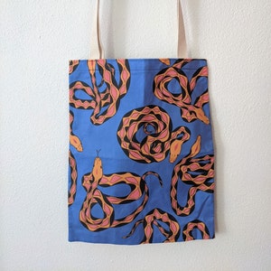 May include: A blue tote bag with a repeating pattern of stylized snakes in orange, pink, and black. The bag has beige straps and is hanging on a wall. The bag is made of fabric and is designed for carrying items.