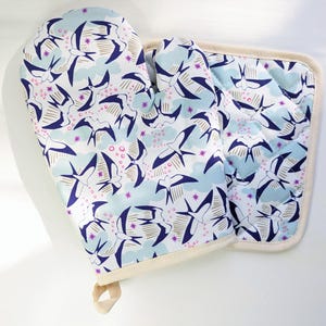 Puede incluir: Un juego de manopla y agarradera a juego. El juego presenta un fondo azul claro con un patrón repetido de pájaros estilizados en azul marino y dorado, con detalles en rosa. Los bordes están ribeteados con una tela color crema.