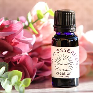 May include: A small, dark purple glass bottle with a black cap, labeled "Devi Essentials 4th chakra creation 1/2 oz (15ml)". The bottle is sitting on a wooden surface with pink flowers in the background.