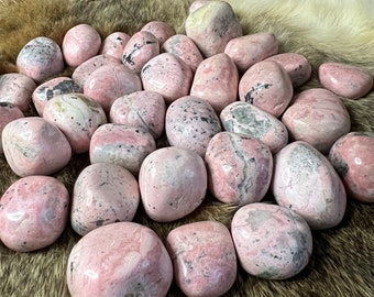 Può includere: Una collezione di pietre rotonde rosa e bianche con una varietà di motivi e texture. Le pietre sono disposte su una superficie di pelliccia sintetica marrone.