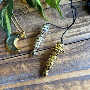 May include: Two metal pendulums, one silver-toned and one gold-toned, each featuring a tiered cylindrical body and a pointed tip, suspended by black cords. Beside them lies a crescent moon pendant with a gold-toned edge and a green stone center.