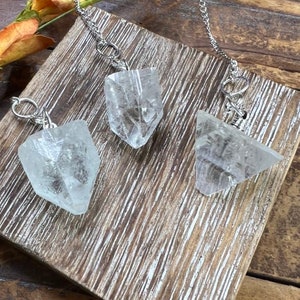 May include: Three clear, faceted crystal pendants, each attached to a silver-toned metal chain link. The crystals have irregular, geometric shapes with visible internal fractures and reflective surfaces, arranged on a textured wooden surface.