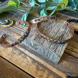 May include: Three copper bracelets with colorful gemstones are displayed on a wooden surface. The bracelets have a hammered texture and are adorned with various colored stones. A gold necklace with a green pendant and black beads are also visible.