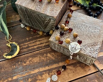 May include: A rosary necklace featuring alternating light brown wooden beads and translucent amber-colored beads linked by silver-tone metal wire. The piece includes a silver-tone medal depicting a dove and a pendant shaped like a pair of angel wings.