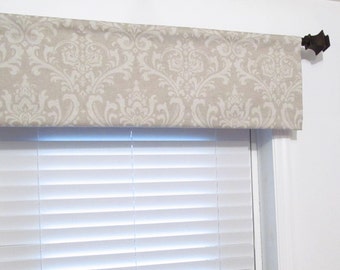 May include: A beige valance with a white damask pattern hangs on a window with white blinds. The valance is attached to a brown metal rod.