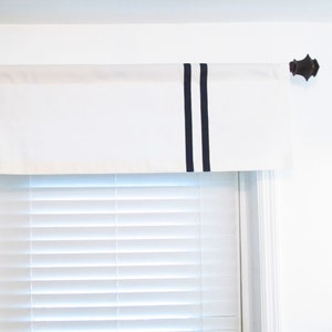 May include: White valance with two black stripes on the right side, hanging on a window with white blinds.