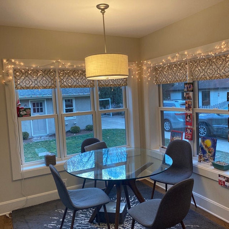 May include: A glass dining table with four chairs in a room with windows and string lights. The table has a glass top and a wooden base. The chairs are upholstered in a dark gray fabric. The windows are decorated with white string lights and have a view of a backyard.