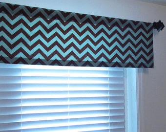 May include: A window valance featuring a repeating chevron pattern in dark brown and light blue. The fabric is suspended from a dark-colored curtain rod with decorative finials, positioned directly above white horizontal window blinds.