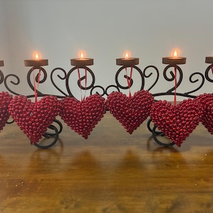 May include: A black metal candle holder featuring six circular platforms for tea light candles. Each platform is adorned with a hanging red heart ornament covered in small, reflective beads, suspended by a thin red ribbon from the metal frame.