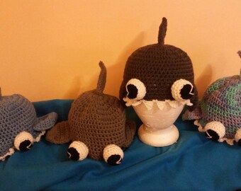 May include: Four crocheted shark hats in different colors, including blue, gray, brown, and purple. Each hat has white eyes and a white mouth.