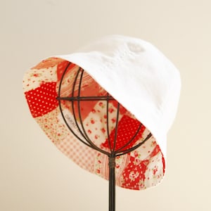 May include: A white bucket hat with a red, pink, and white patchwork lining. The lining is made of different fabrics with floral and geometric patterns.