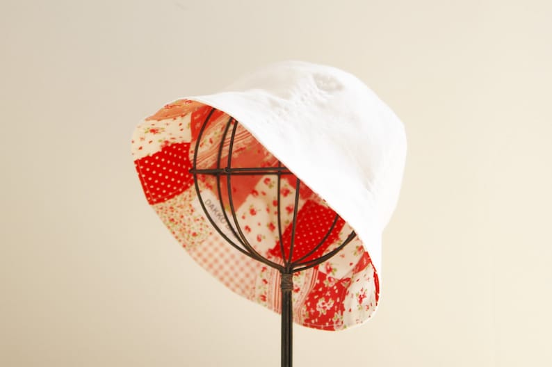 May include: A white bucket hat with a red, pink, and white patchwork lining. The lining is made of different fabrics with floral and geometric patterns.