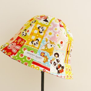 Puede incluir: Un sombrero cubo de parches amarillo, rojo, verde y rosa con estampado de panda y conejo. El sombrero tiene una etiqueta que dice "Dakko Baby".