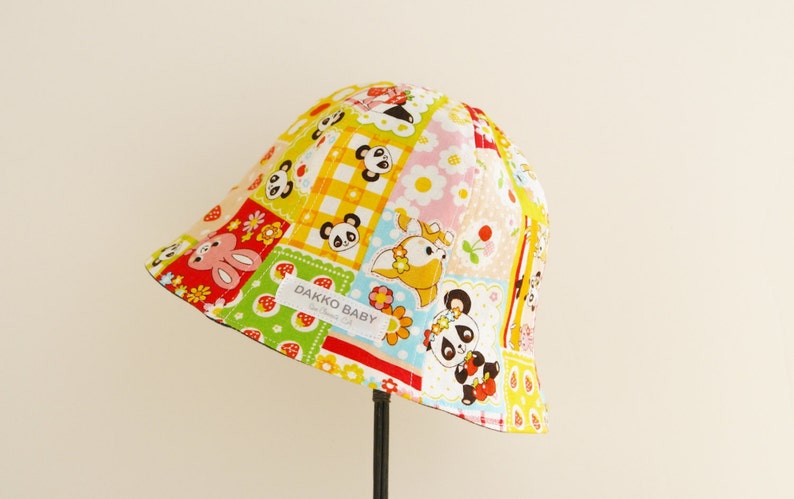 Puede incluir: Un sombrero cubo de parches amarillo, rojo, verde y rosa con estampado de panda y conejo. El sombrero tiene una etiqueta que dice "Dakko Baby".