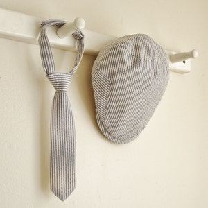 May include: A striped tie and a striped cap hang on a white wooden rack. Both items are made of a light-colored fabric with thin, dark stripes.