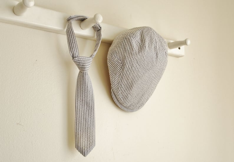 May include: A striped tie and a striped cap hang on a white wooden rack. Both items are made of a light-colored fabric with thin, dark stripes.