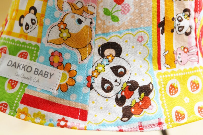 Puede incluir: Una tela colorida con un dise&ntilde;o de patchwork que presenta osos panda, un ciervo, flores y fresas. La tela tiene un fondo blanco con un dise&ntilde;o de patchwork azul, amarillo, rojo y verde. La tela tiene el texto "DAKKO BABY San Clemente, CA" en una etiqueta blanca.