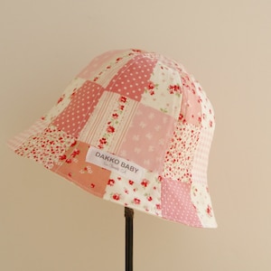 May include: A pink and white patchwork bucket hat with a floral pattern. The hat has a label that says "Dakko Baby".
