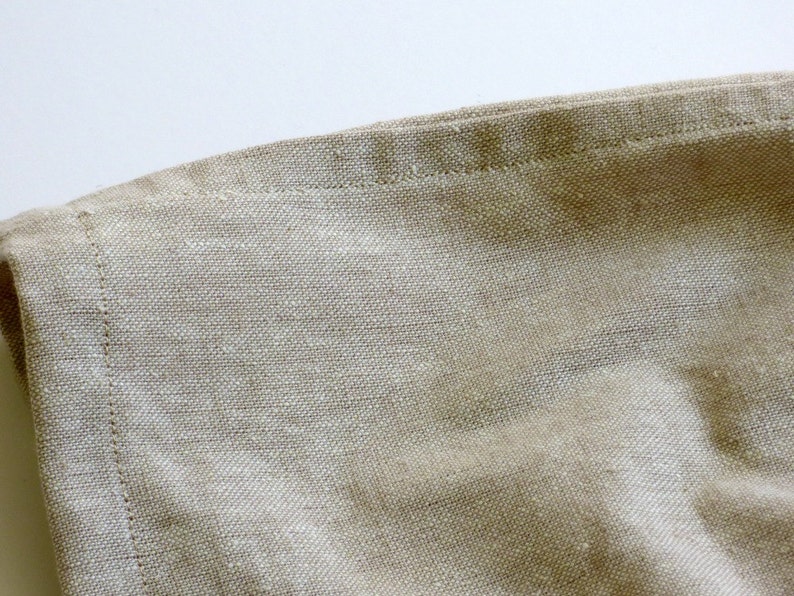 May include: Close-up of a beige linen fabric with a stitched edge. The fabric has a natural, textured look and feel.